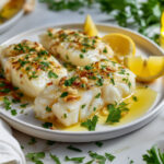 Filets d’aiglefin dorés au four, nappés d’huile d’olive, parsemés d’ail et de persil, servis avec quartiers de citron – recette d’aiglefin à l’ail.