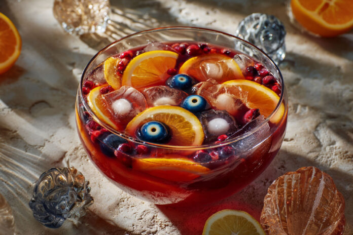 Grand saladier de punch d’Halloween rouge sang, garni de rondelles d’agrumes, fruits rouges et d’une main glacée, prêt à être servi.