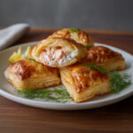 Feuilletés dorés au saumon et fromage frais citronné, garnis d’aneth, sur une assiette accompagnée de salade verte – recette savoureuse et facile.