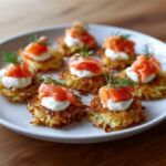 Mini-röstis dorés à la poêle, garnis de fromage frais, saumon fumé et brins d’aneth, présentés sur une assiette blanche.